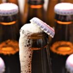 Botella de cerveza abierta rebosando espuma junto a otras botellas selladas